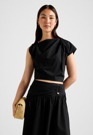 Kobieta ubrana w czarny bezrękawnik i spódnicę z marszczonym detalem w talii, trzyma beżową plecioną kopertówkę na jednolitym tle.