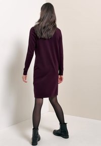 Femme en robe longue manches longues violet foncé jusqu'aux genoux, collants noirs transparents et bottines noires à lacets, debout contre un mur clair uni.
