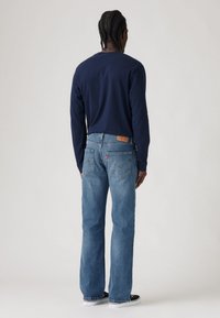 Camisa de manga larga azul marino combinada con jeans de mezclilla azul claro. Los jeans presentan un parche de cuero con el logo y la típica costura de los bolsillos traseros. Calzado negro visible.