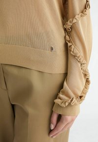 Maglia beige a maniche lunghe con un morbido knit testurizzato, dettagli arricciati lungo le maniche e un orlo a coste con un piccolo dettaglio metallico.