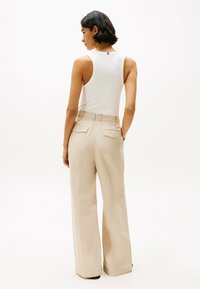Pantaloni beige a gamba larga con un tessuto testurizzato, dotati di tasche posteriori e vita con cintura, abbinati a un top bianco aderente senza maniche.