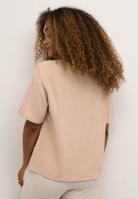 Kortärmad, ljusbeige blus med avslappnad passform. Slät textur och subtilt vävt mönster. Sömmen böjer sig lätt i ryggen.