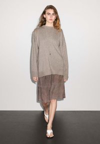 Übergroßer beiger Strickpullover kombiniert mit einem knielangen braunen Wildlederrock, ergänzt durch weiße flache Sandalen. Einfaches Detail mit einer silbernen Halskette.