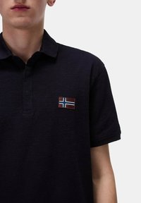 Polo shirt blu scuro con colletto in tessuto testurizzato e un piccolo patch ricamato della bandiera norvegese sul lato sinistro.