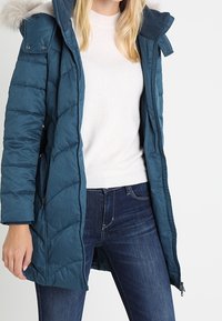 Teal pufferjakke med hætte trimmet med pels, har quiltet design, lynlås-lommer og en længde til midt på låret. Bæres over en let sweater.