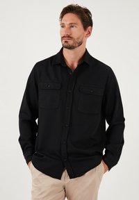 Cămașă neagră cu mânecă lungă din bumbac, cu guler tip button-down, având două buzunare pe piept și un tiv drept. Purtați-o cu pantaloni bej.
