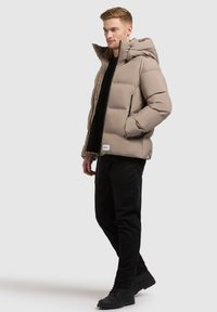Pufferjack in licht beige, met een ritsopening aan de voorkant, een capuchon, twee zijzakken en elastische manchetten. Gdragend met zwarte broek en schoenen.