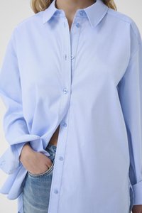 Camisa de algodón azul claro con botones, que presenta un cuello clásico, mangas largas y un ajuste holgado. Los detalles notables incluyen botones azules.