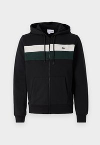 Crna zip-up dukserica od mekog materijala. Ima bijelu i tamnozelenu horizontalnu prugu preko grudi i prednji džep. Kapuljača je uključena.