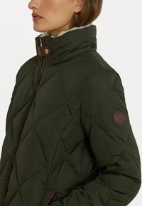 Tmavozelená prešívaná bunda s vysokým golierom, hnedými koženými detailmi a okrúhlou logom na rukáve. Mäkčená podšívka viditeľná na golieri.