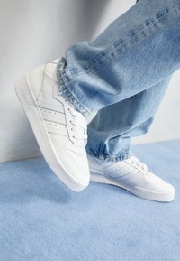 Person, der weiße Adidas-Sneaker mit drei Streifen trägt und helle blaue Jeans hat, steht auf einem hellblauen Teppich.