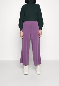 Maglione a coste verde scuro e pantaloni larghi viola chiaro. I pantaloni hanno un elastico in vita. Sneakers bianche dalla texture chiara completano l'outfit.