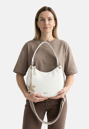 Femme portant une chemise et un pantalon marron, tenant un sac blanc avec un porte-clés étoile de mer et une bandoulière, sur un fond uni.