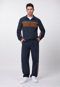 Άνδρας στέκεται με τα χέρια στις τσέπες, φορώντας σκούρο μπλε φόρμα με καφέ ρίγα στο στήθος και τις πλευρές, και λευκά αθλητικά παπούτσια, ουδέτερο φόντο.