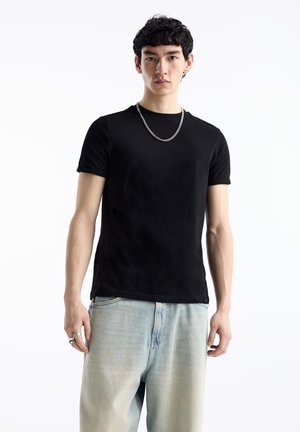 Tricou din bumbac negru, cu mâneci scurte, guler rotund, purtat cu blugi denim de un albastru deschis, ușor decolorat. Accesoriu: colier din lanț argintiu.