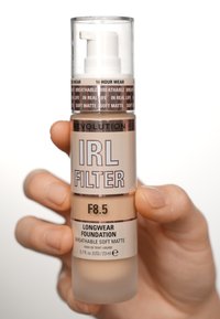 Bouteille pompe transparente contenant un fond de teint beige clair. L'étiquette indique "FILTRE IRL F8.5", "FOND DE TEINT LONGUE DURÉE", et "MATTE SOUPLE RESPIRANT".