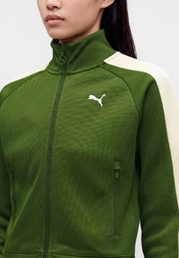 Γυναίκα που φοράει πράσινο μπουφάν Puma με φερμουάρ, με λευκές ρίγες στους ώμους και λευκό λογότυπο Puma στο στήθος.