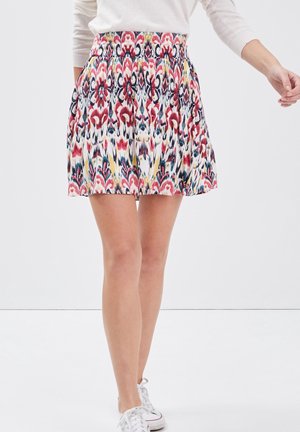 Femme portant une mini-jupe taille haute colorée avec un motif abstrait et des baskets blanches, marchant sur un fond blanc uni.