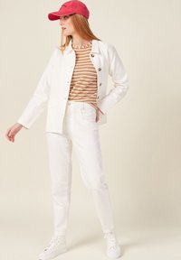 Giacca di jeans bianca con bottoni, top a righe arancioni e bianche, jeans bianchi, cappellino rosso e sneakers bianche. Outfit casual con linee pulite.