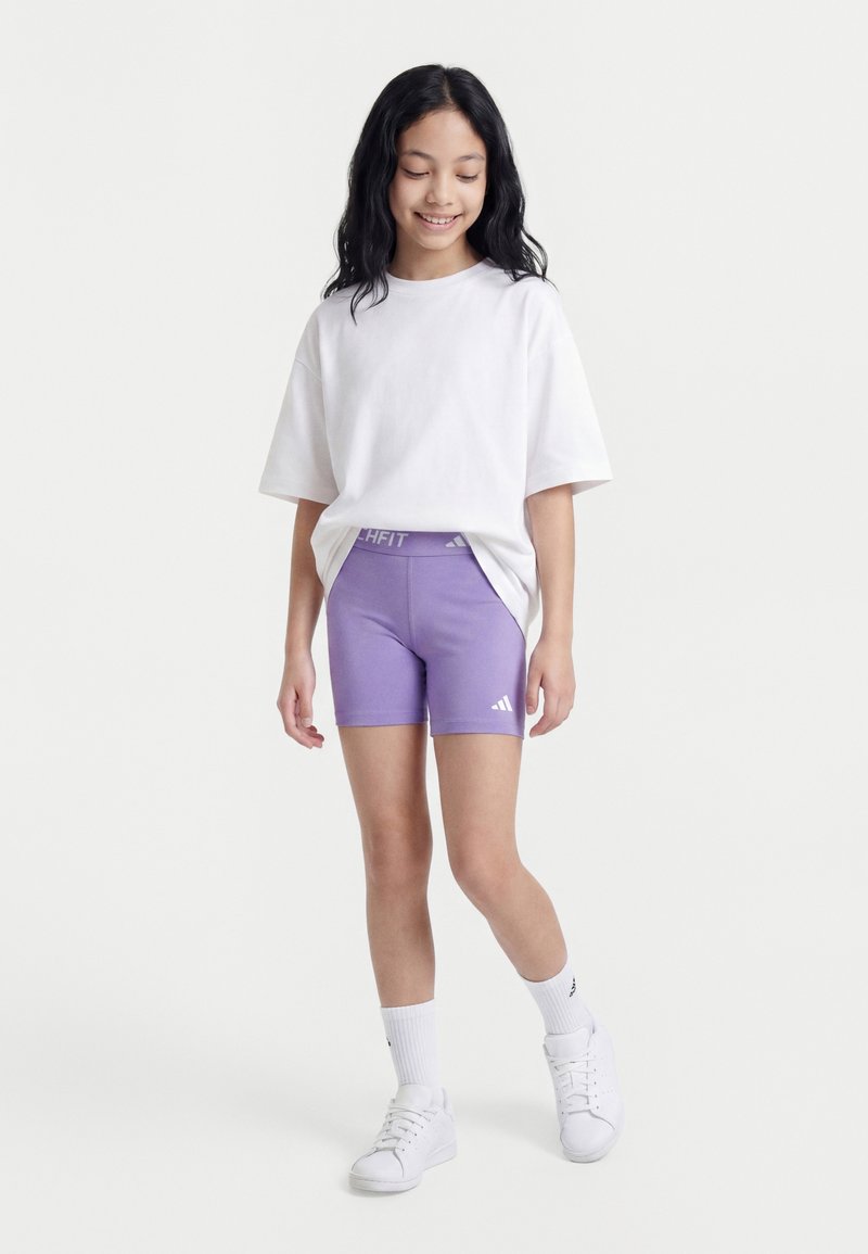 Fille debout, portant un t-shirt blanc oversize, un short de sport violet, des chaussettes blanches et des baskets blanches, souriante et regardant vers le bas.