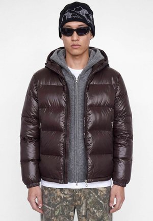 Un mannequin se tient devant un fond blanc, vêtu d’une doudoune marron foncé, d’un sweat à capuche gris, d’un pantalon à motifs, de lunettes de soleil et d’un bonnet noir.