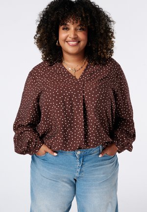 Femme souriante aux cheveux bouclés portant un chemisier marron à pois et un jean bleu clair, les mains dans les poches, sur un fond uni.