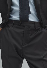 Schwarze, maßgeschneiderte Trousers mit glatter Textur, gerader Schnitt und einem Einzelknopfverschluss, kombiniert mit einem blauen gestreiften Hemd und einer dunklen Strickkrawatte.