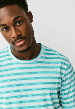 Young man with short curly hair and facial hair wearing a turquoise and white striped T-shirt, tilting head slightly against a plain background.