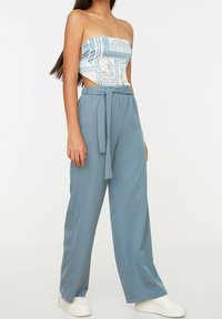 Světlomodré kalhoty s širokými nohavicemi a uvázaným páskem, vyrobené z měkké látky. V kombinaci s bílým vzorovaným topem bez ramínek.