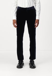 Námornícky sametové nohavice so slim fit, s hladkou textúrou a jemným leskom, v kombinácii s čiernymi koženými topánkami a bielou košeľou.