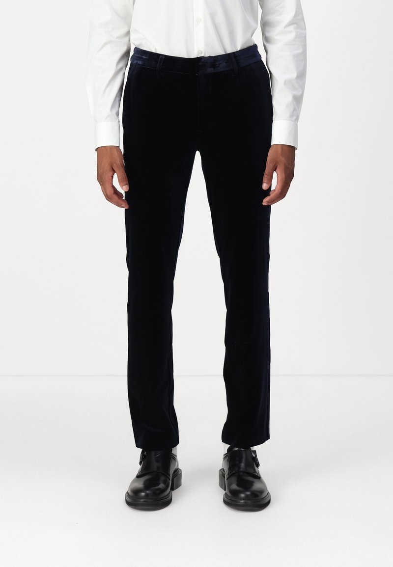 Námornícky sametové nohavice so slim fit, s hladkou textúrou a jemným leskom, v kombinácii s čiernymi koženými topánkami a bielou košeľou.