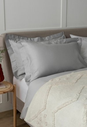 Beiges gepolstertes Bett mit zwei grauen Satin-Kissen, einer cremefarbenen strukturierten Decke und einem hölzernen Nachttisch mit einer roten Vase und Grünpflanzen.