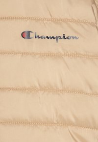 Smėlio spalvos pūkinė striukė su siuvinėtu paviršiumi. Priekyje yra ryškus metalinis „Champion“ logotipas mėlynos ir raudonos spalvos.