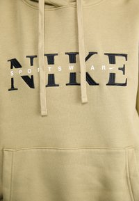 Olive spalvos džemperis su kengūros kišene, apdailintas juodais išsiuvinėtais "NIKE" rašmenimis ir mažesniu "SPORTSWEAR" tekstu baltai.