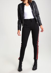 Mujer con chaqueta de cuero negra, camiseta blanca, pantalones negros con franjas laterales rojas y blancas, y botas de tobillo negras con cordones, de pie frente a un fondo blanco.