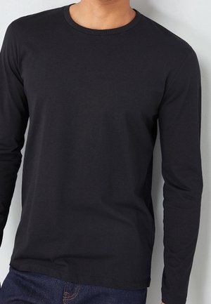 Personne portant un t-shirt noir uni à manches longues et un jean bleu, debout contre un mur clair.