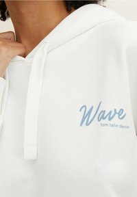 Persona con una sudadera blanca con cordones, que presenta el texto azul claro "Wave tom tailor denim" en el pecho.