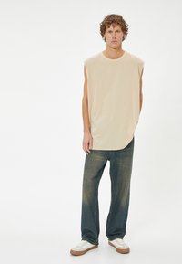 Beige mouwloze T-shirt met een ronde hals, gecombineerd met blauwe hooggetailleerde jeans en witte sneakers met bruine accenten. Gladde stof textuur.