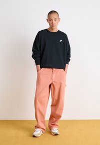 Tengerészkék pamut pulóver fehér Nike logóval, lazán illeszkedő, világos rózsaszín nadrággal párosítva. A lábbeli fehér alapszínű, több színnel díszítve.