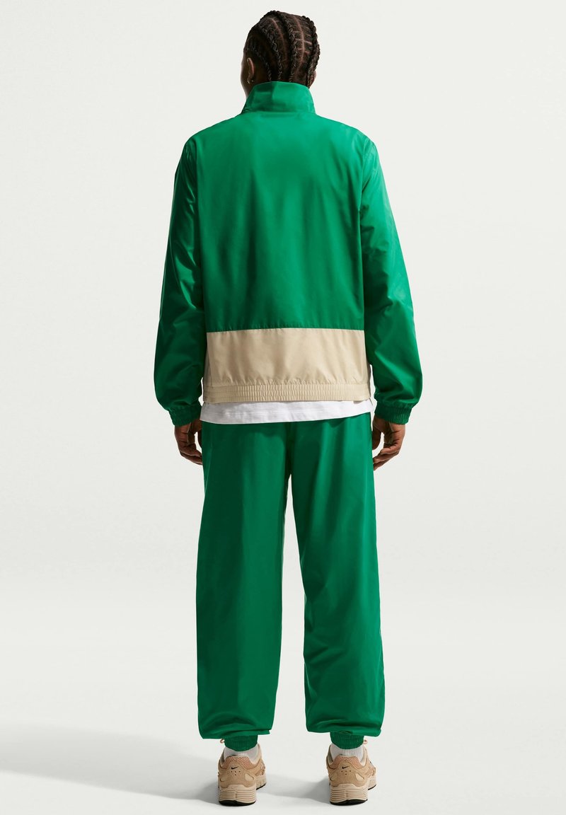 Person mit geflochtenem Haar, die eine grün-beige Jacke, grüne Hose, weißes Hemd und beige Sneakers trägt, steht mit dem Rücken zur Kamera vor einem einfarbigen Hintergrund.