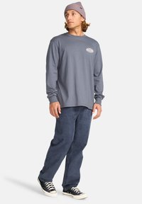 Grau langärmliges Shirt mit einem Logo auf der linken Brust, kombiniert mit blauen Cordhosen und schwarzen Sneakers mit weißen Akzenten.