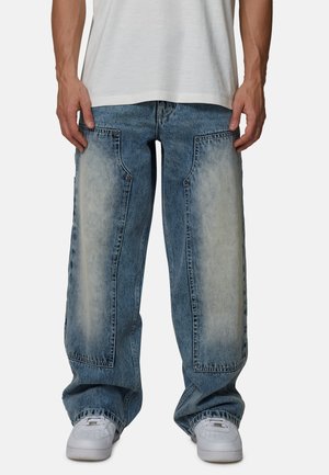 Wijdvallende spijkerbroek met lichte, vervaagde blauwe stukken, zichtbare stiksels en twee voorzakken. Gecombineerd met witte sneakers.