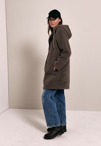 Femme portant une casquette noire, une veste polaire grise à capuche, un jean bleu et des bottes noires, debout de profil, les mains dans les poches de la veste, devant un fond uni.