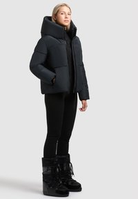 Svart pufferjacka med hög krage och boxig passform, bärs över svarta leggings och höga snörkängor med glänsande finish.
