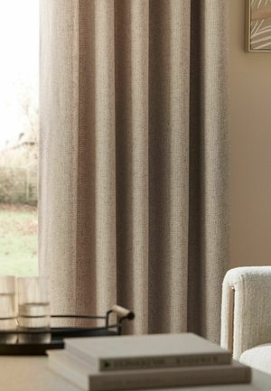 Beigefarbener, strukturierter Vorhang, der an einem Fenster mit unscharfem Außenblick hängt, ein hellfarbiger Sessel und ein Tisch mit Büchern und Brillen.