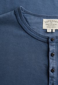 Zbliżenie na dekolt niebieskiej bawełnianej koszuli z trzema czarnymi guzikami i białą metką Superdry.