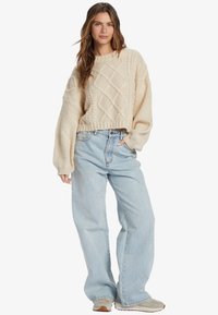 Pull en tricot crème à mailles torsadées avec des manches longues et volumineuses ; associé à un jean large taille haute bleu clair et des baskets beiges.