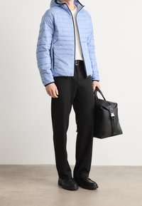 Ljust blå puffjacka med huva, svarta dragkedjedetaljer, bärs med svarta byxor och en strukturerad svart väska, mot en neutral bakgrund.