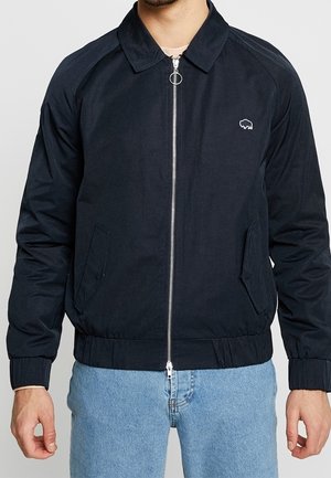 Homme portant une veste noire zippée avec col et poches latérales, associée à un jean bleu clair, montrée du cou aux hanches.