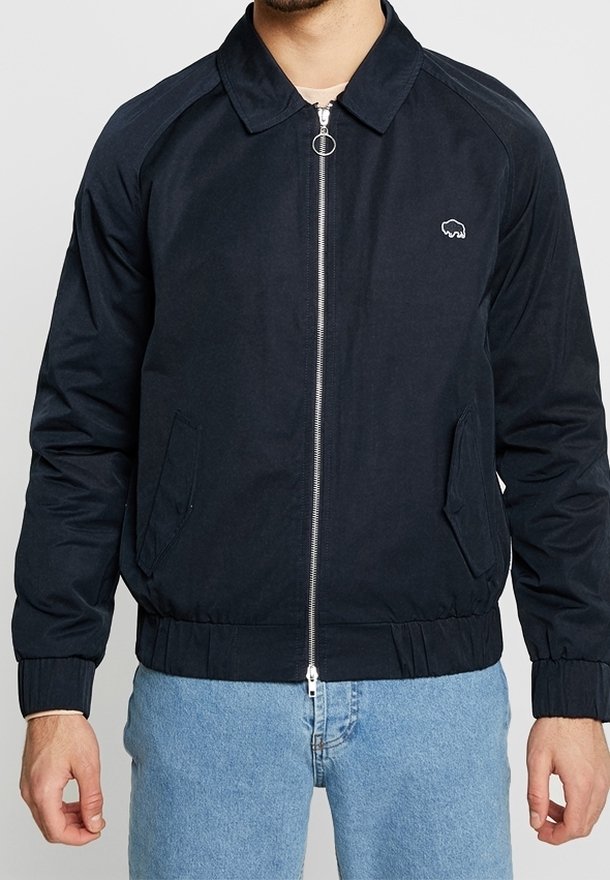 Homme portant une veste noire zippée avec col et poches latérales, associée à un jean bleu clair, montrée du cou aux hanches.